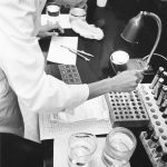 A black and white photo of a woman in a lab coat working with vials and pipettes in a lab.