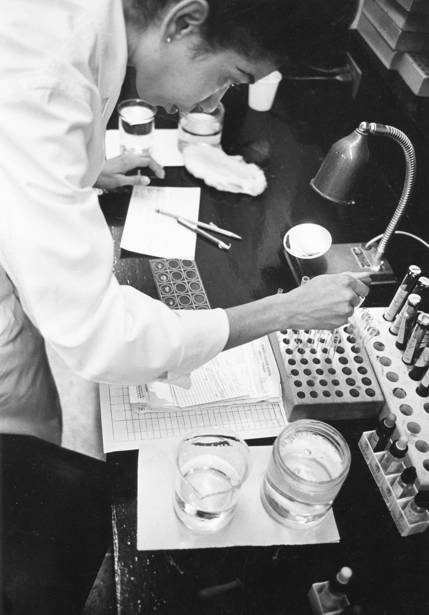 A black and white photo of a woman in a lab coat working with vials and pipettes in a lab.