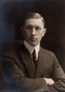 A black and white photo of a man dressed in a suit. He wears round spectacles.