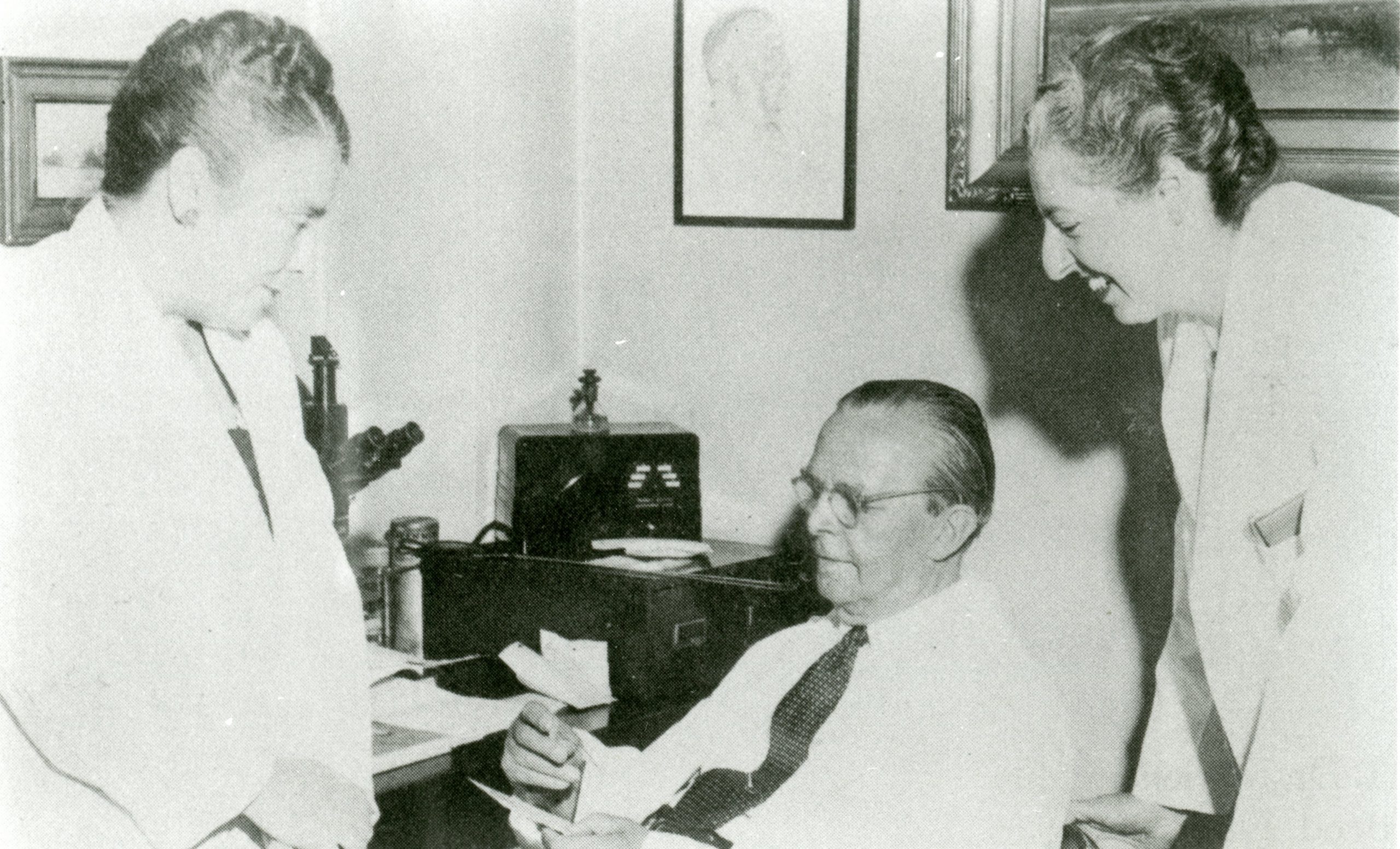 A black and white photo of two women in lab-coats speaking with an older man in a dress shirt and tie.