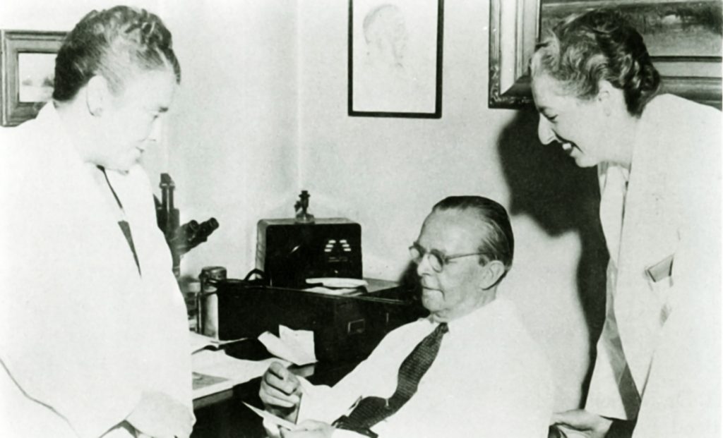 A black and white photo of two women in lab-coats speaking with an older man in a dress shirt and tie.
