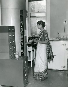 A black and white photo of a young woman wearing a sari acessing a medical supply cupboard.