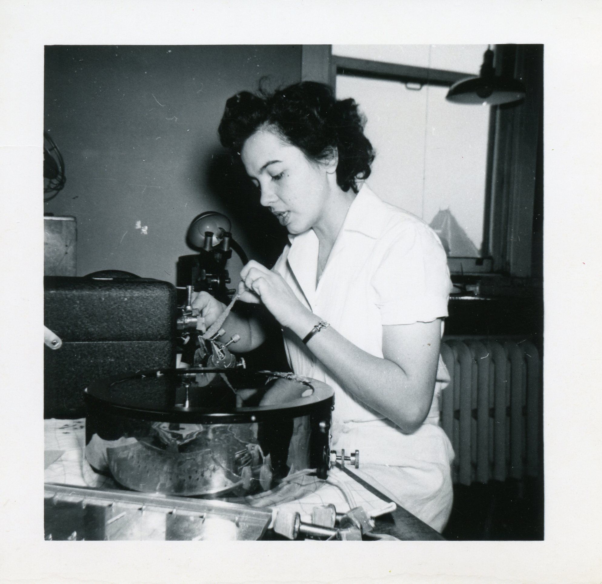 A black and white photo of a woman in a lab-coat checks on scientific equipment.