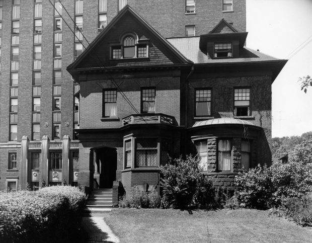 A black and white photo of a two story house. In the background looms the hospital.