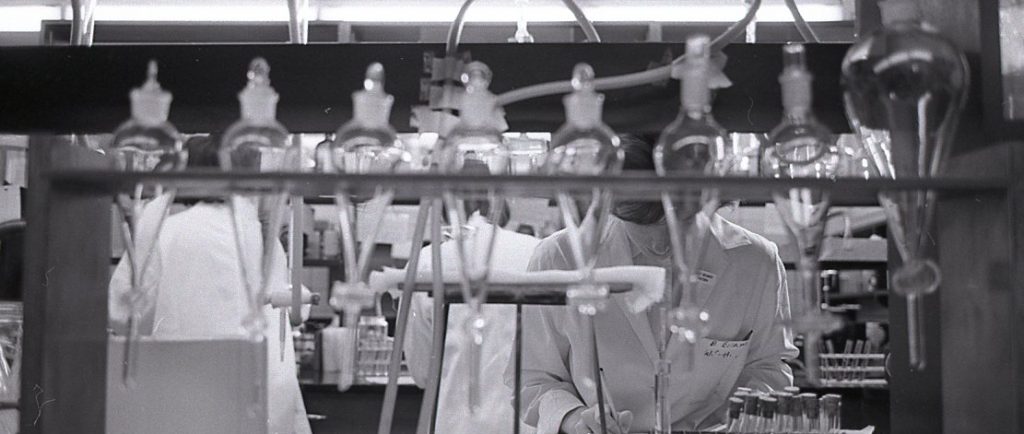 A black and white photo of a three women in lab coats are visible behind a row of beakers and scientific equipment.