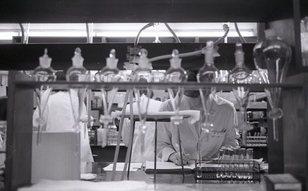 A black and white photo of a three women in lab coats are visible behind a row of beakers and scientific equipment.