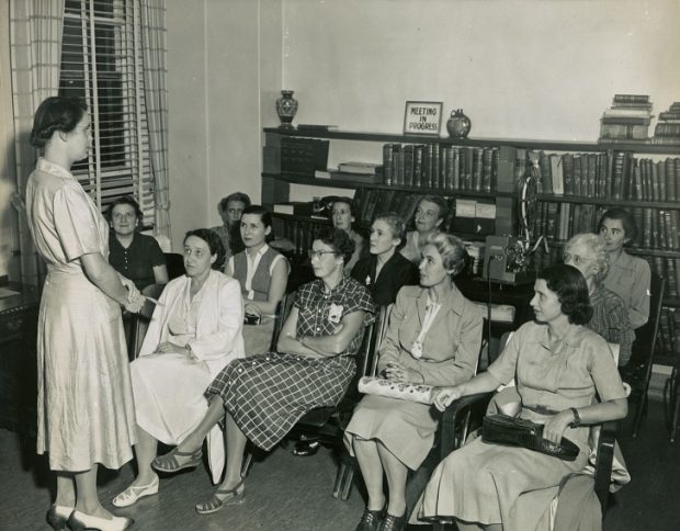 A black and white photo of a group of seated women listening to a woman speaking. They are in a room lined with books.