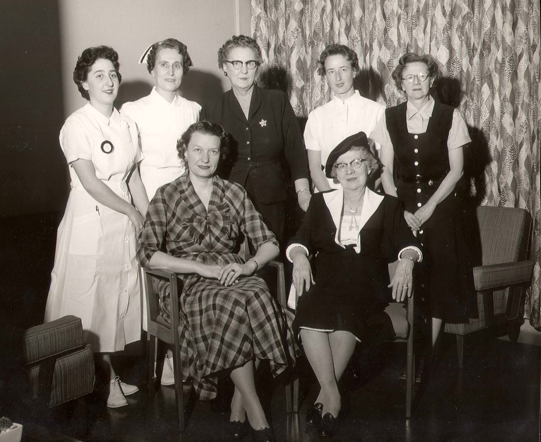 A black and white photo of seven ladies posing. Two are seated, and a number of those standing are wearing nurse uniforms.