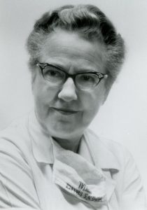 A black and white portrait of a woman in glasses. A medical mask hangs on her neck.