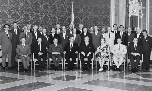 A black and white photo of a group of formally dressed people. Of the thirty or so, one is a woman. They pose in an elegant room.