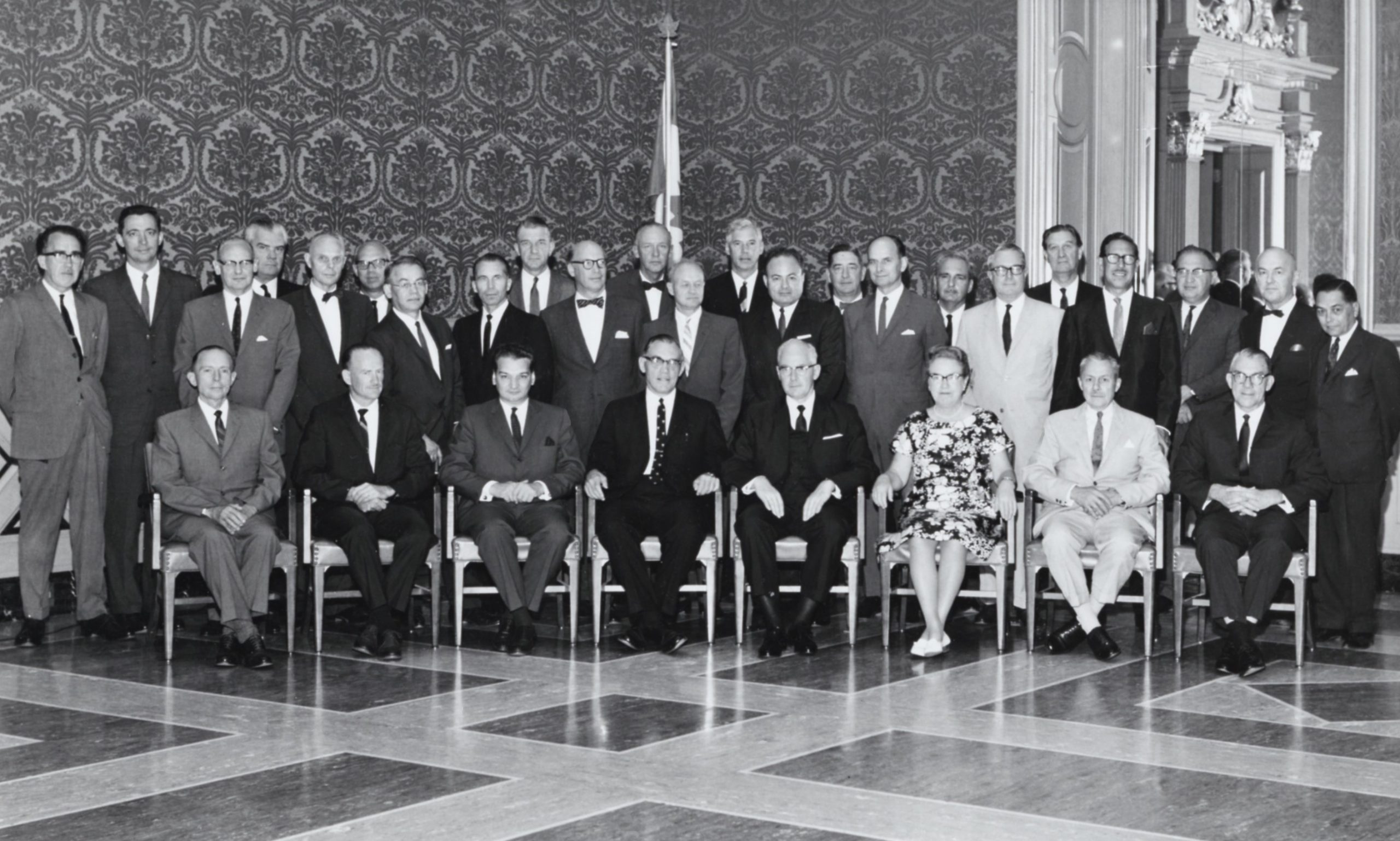 A black and white photo of a group of formally dressed people. Of the thirty or so, one is a woman. They pose in an elegant room.