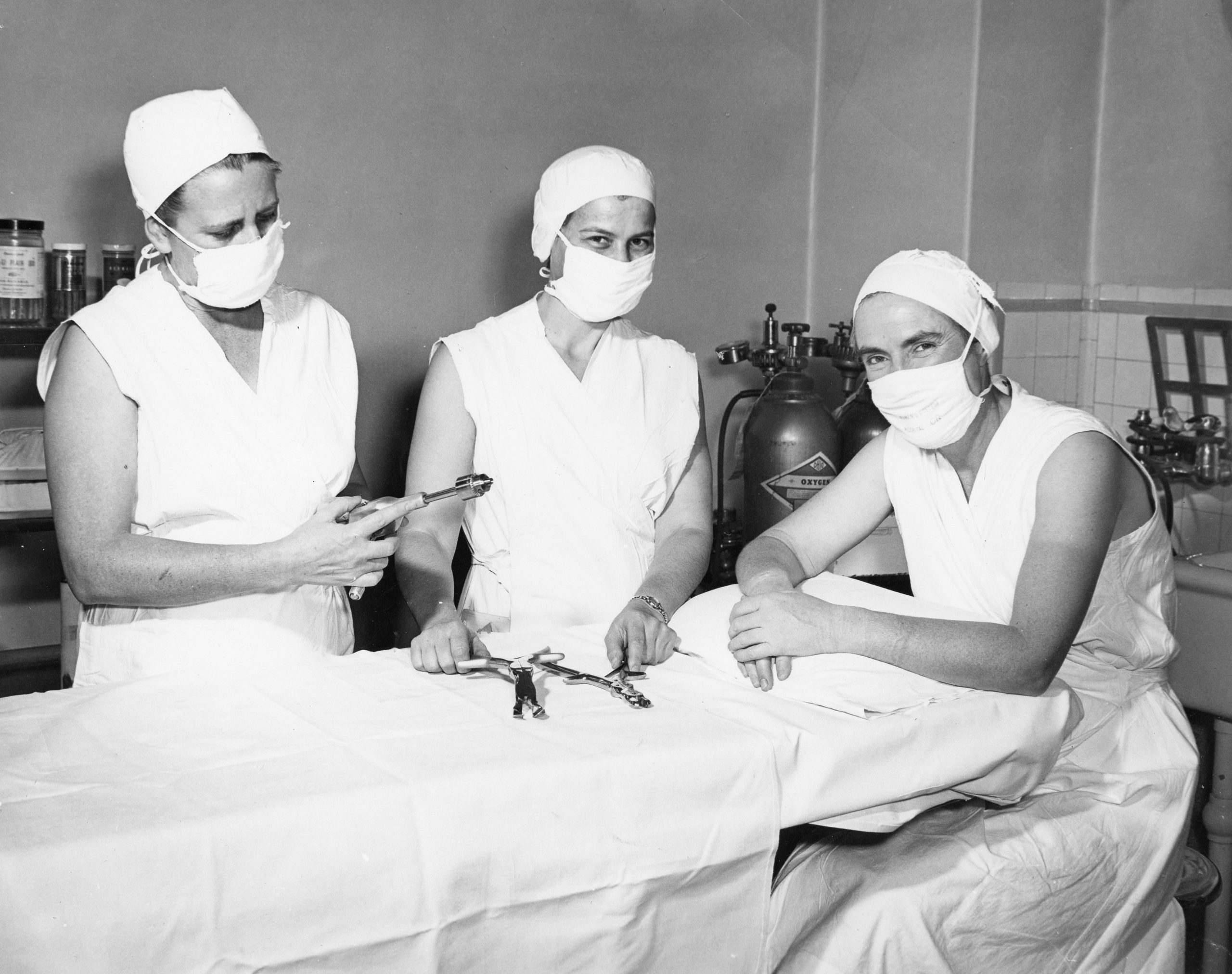 A black and white photo of three people in scrubs, posing by an operating table. Their eyes are smiling.