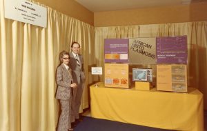 A yellow tinted coloured photo of a man and woman in suits standing by an exhibition table featuring a television and poster boards.