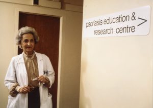 A coloured photo of an older woman wearing a lab coat looking down a hallway by a sign reading 