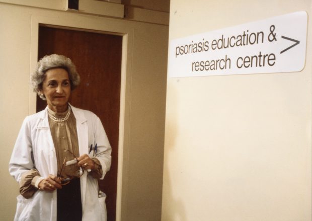A coloured photo of an older woman wearing a lab coat looking down a hallway by a sign reading psoriasis education & research centre.