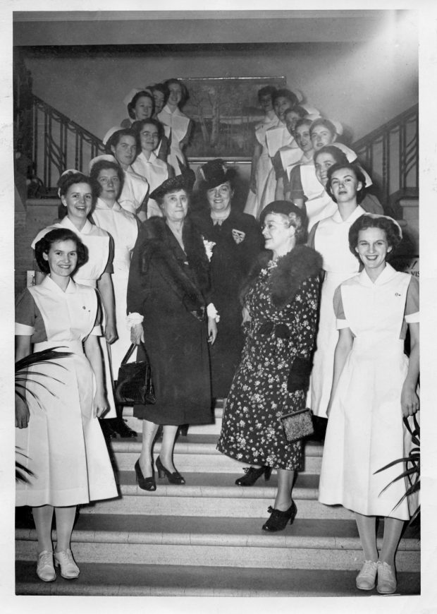 A black and white photo of a group of women on a staircase. Two rows of young women in nurses uniforms line the sides, while three older women, elegantly dressed, stand in the centre.