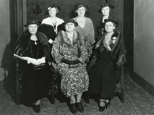 Photo en noir et blanc de six femmes d’âge mûr posant avec des chapeaux et des manteaux élégants.