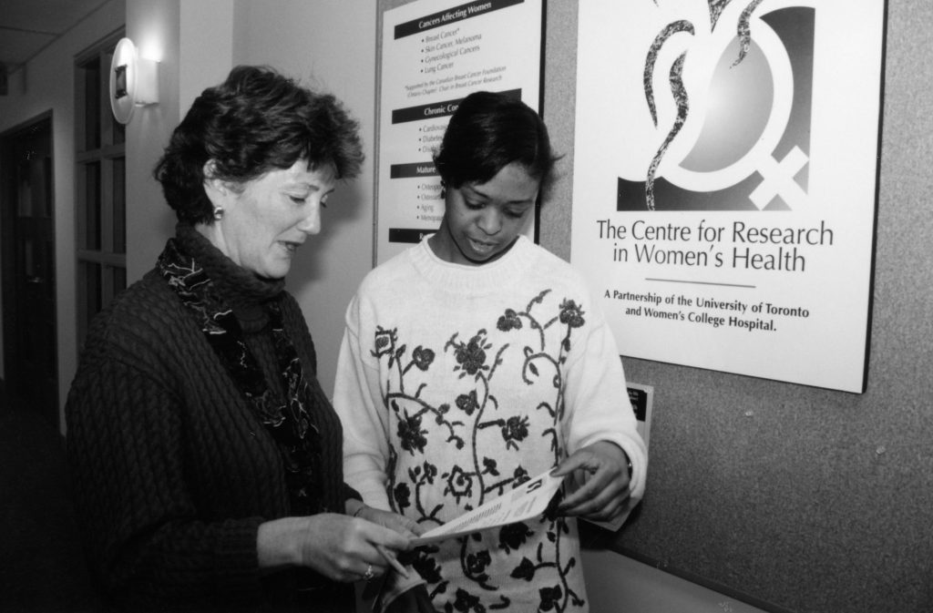 Two women examine a brochure; a large sign on the wall by them reads 