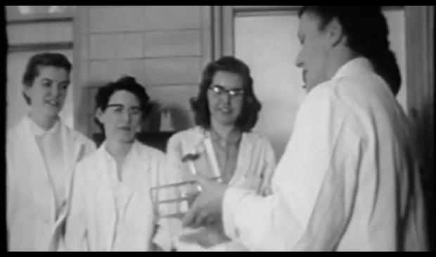 A frame from a black and white video of women in lab coats learning from an older woman.