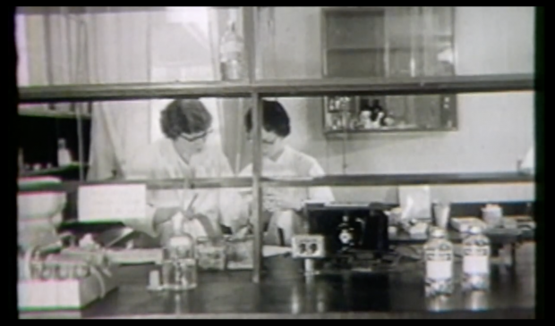 A frame from a black and white video of two women working in a laboratory.