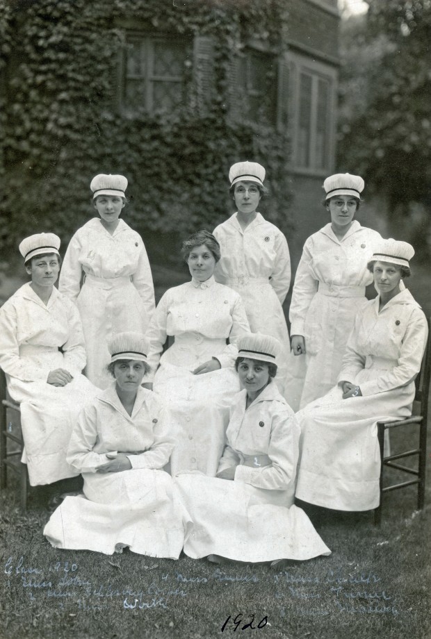 Eight women in full nurses's uniforms stand, sit, and kneel, smiling at the camera.