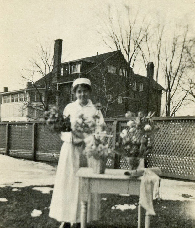 A smiling young woman in a nurse's uniform holds a bouquet. She is outside, and near her a table holds vases and a rolled and ribboned paper.