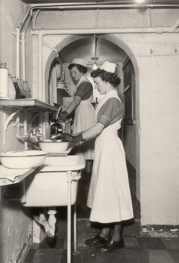 Two women in nurses' uniforms wearing gloves and washing at a series of sinks.