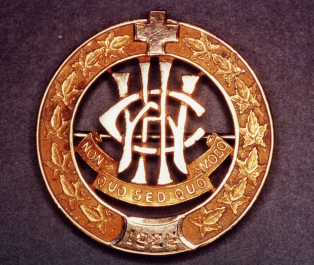 Circular pin, outlined by leaves, topped with a cross. The letters W, C, H, are entwined above the motto.