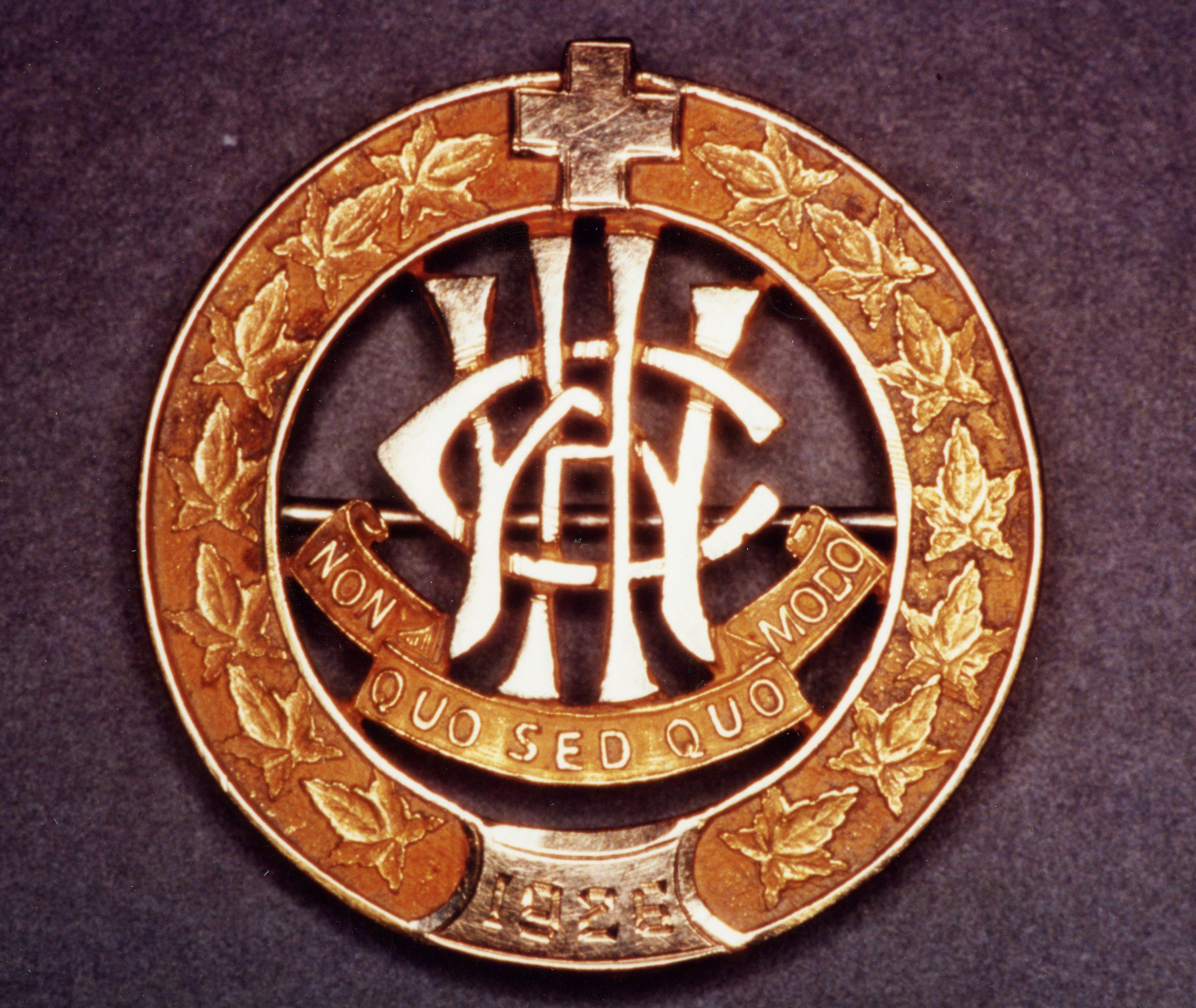 Circular pin, outlined by leaves, topped with a cross. The letters W, C, H, are entwined above the motto.
