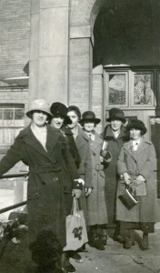 Seven women wearing long coats and hats, before a tall arched doorway.