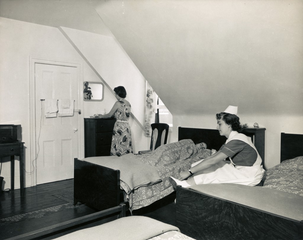 Une femme en robe devant un miroir, une autre en uniforme d’infirmière sur un lit et une troisième endormie. Il y a trois lits dans la chambre.