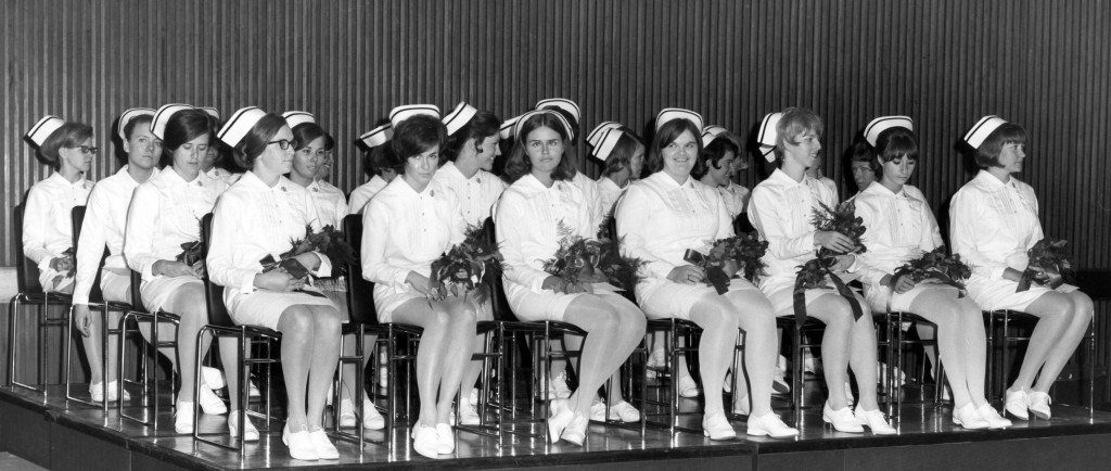 Over 20 women sit on a stage in full nurses' uniforms, holding flowers.