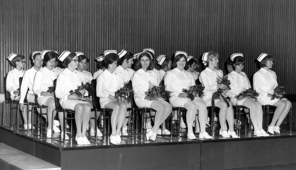 Over 20 women sit on a stage in full nurses' uniforms, holding flowers.