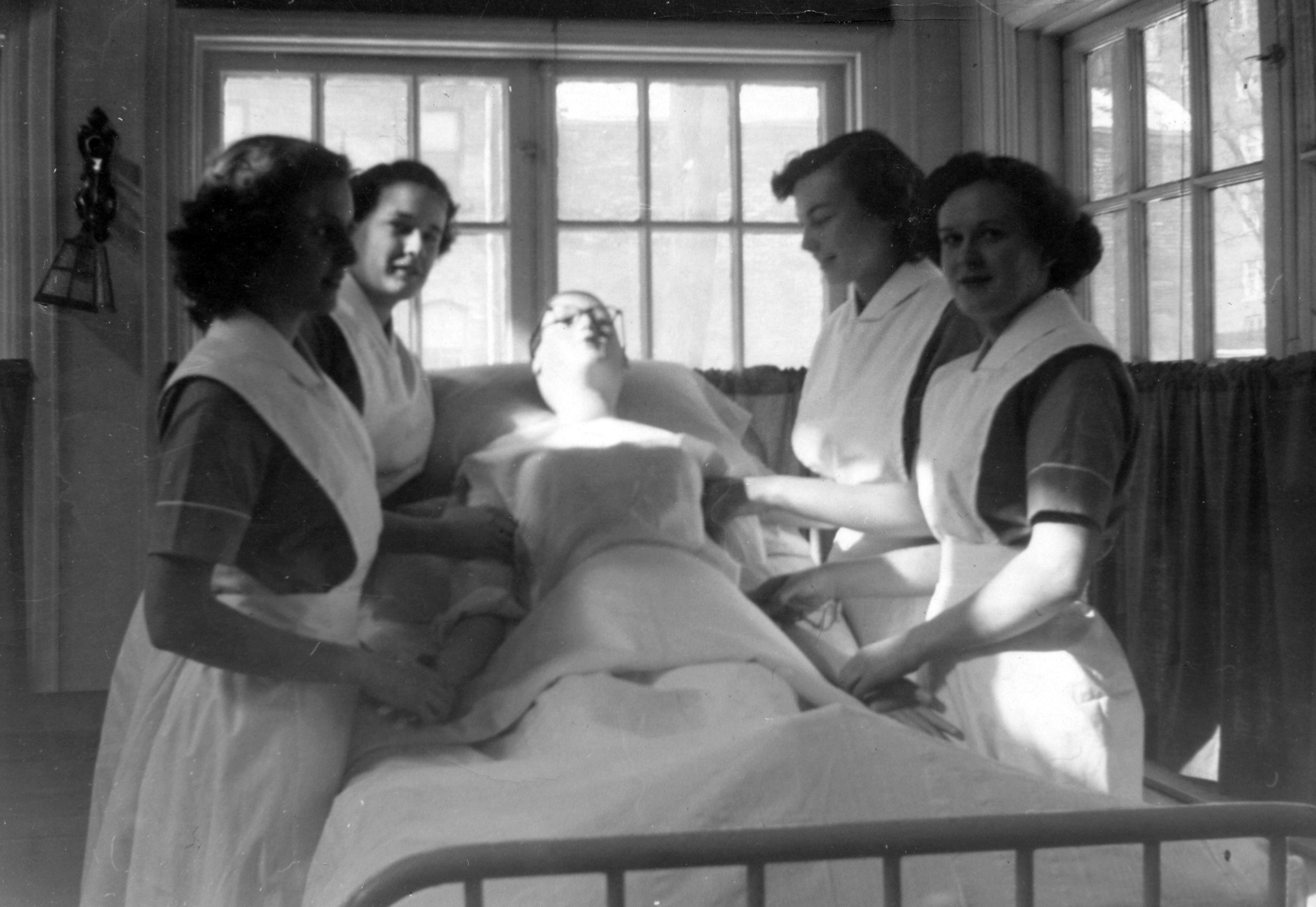 Four women in nurses' uniforms without caps surround a bed holding a mannequin.