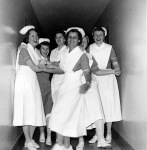 Six laughing and gesturing women in nurses' uniforms crowd a narrow hallway.