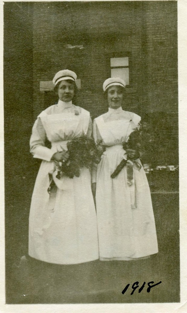 Two women in nurses' uniforms stand before a brick building holding bouquets and smiling.
