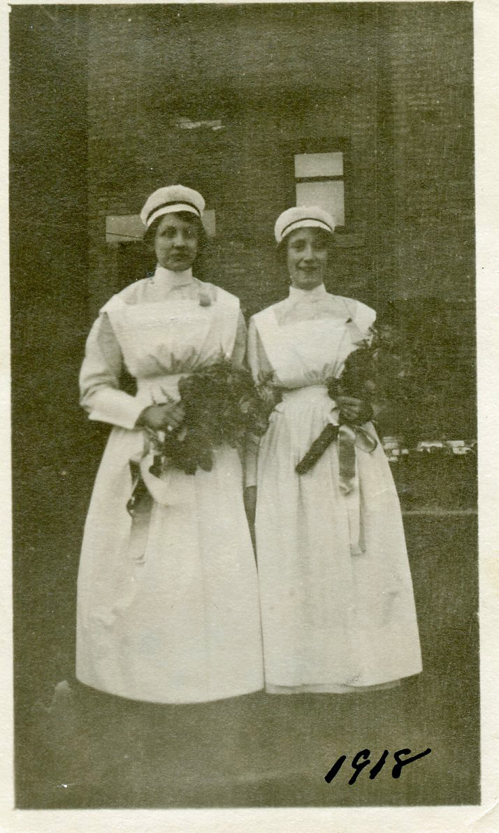 Two women in nurses' uniforms stand before a brick building holding bouquets and smiling.