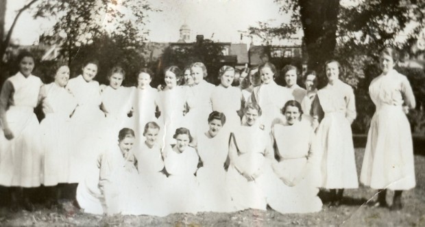 Over 20 smiling woman stand and kneel outside in nurses' uniforms, without caps.