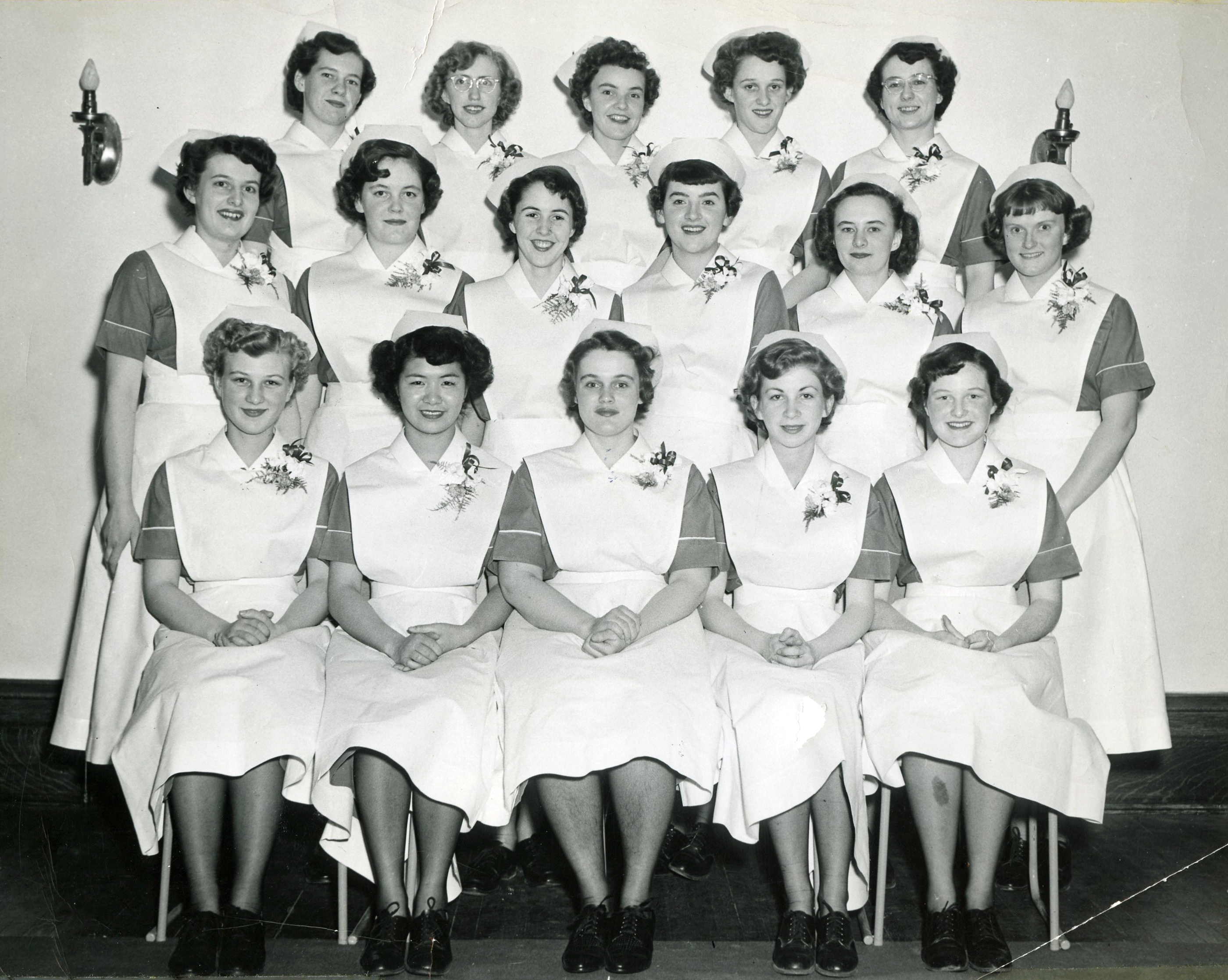 16 smiling women in nurses' uniforms sitting and standing for a formal photo.
