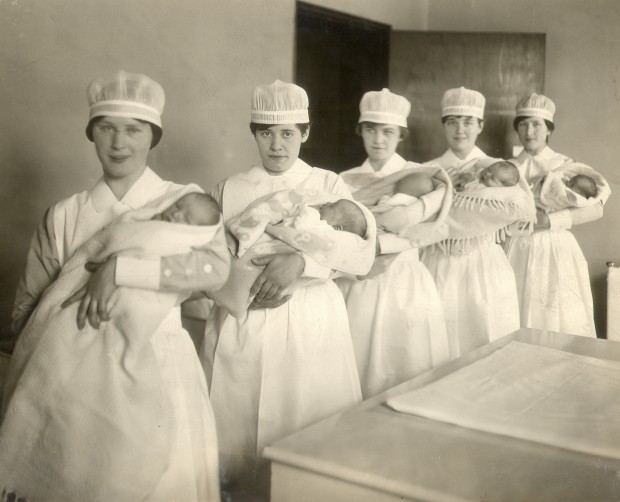 Five young women hold bundled infants and pose for the camera. They are wearing nurses' caps.