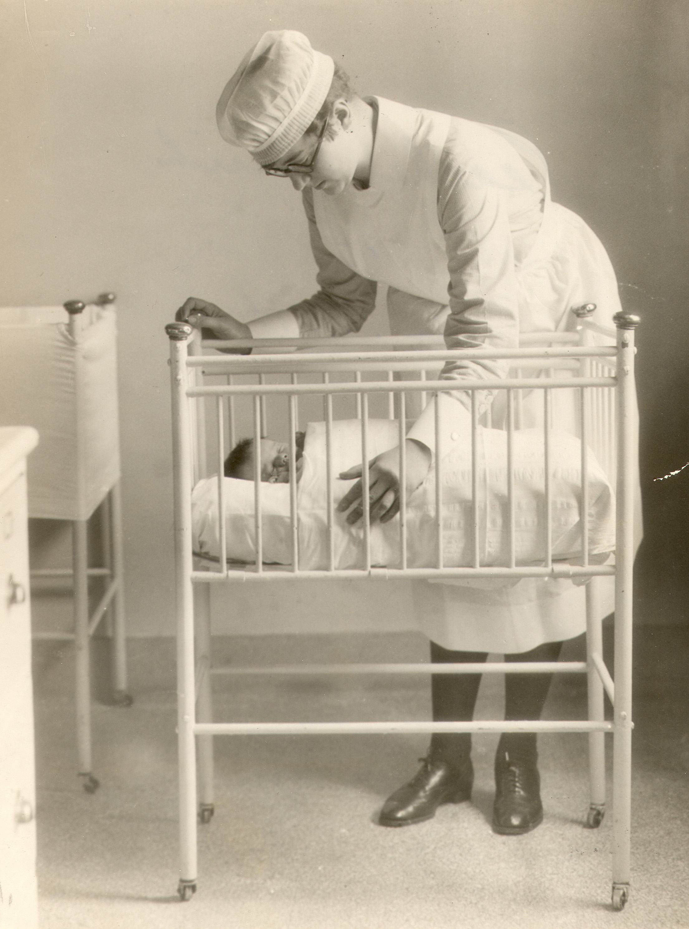 A young woman leans over a crib with a baby in it. She is wearing a nurses' uniform.