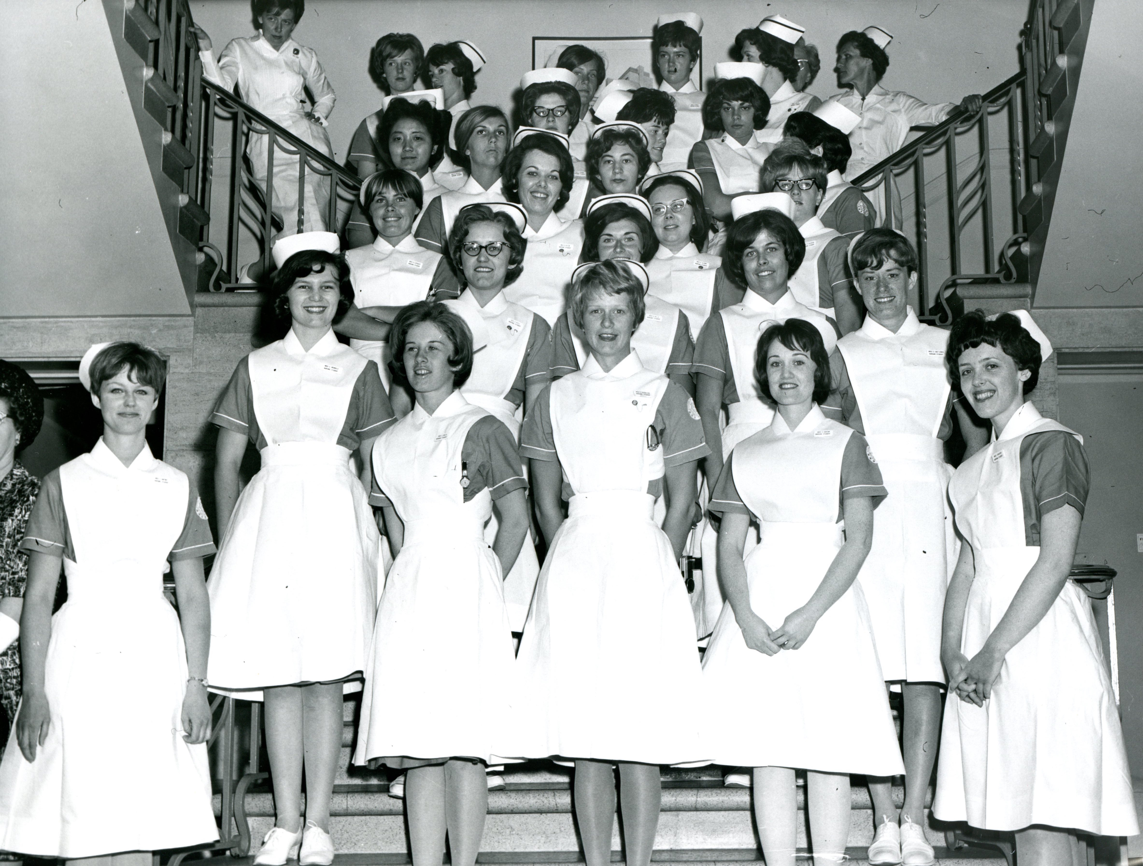 A large group of women pose formally for a photograph on a set of stairs. They are wearing nurses' uniforms with caps.