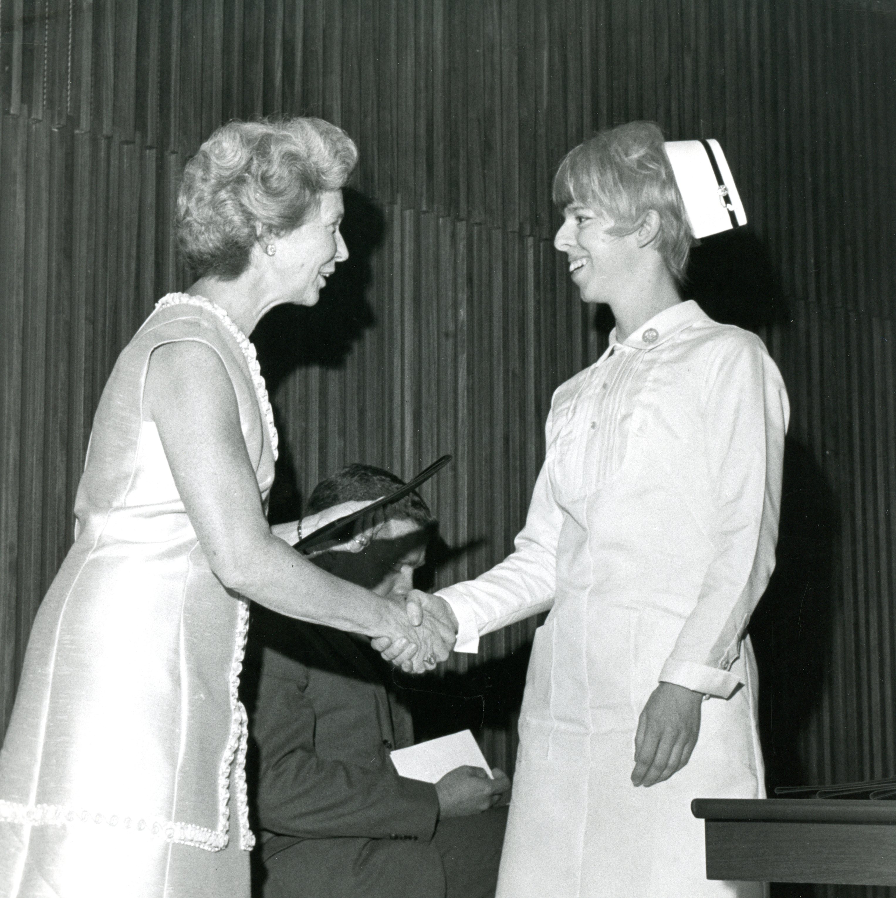 Two women shake hands, smiling. One is dressed in a formal dress, the other in nurses' uniform with cap. A man stands in the background.