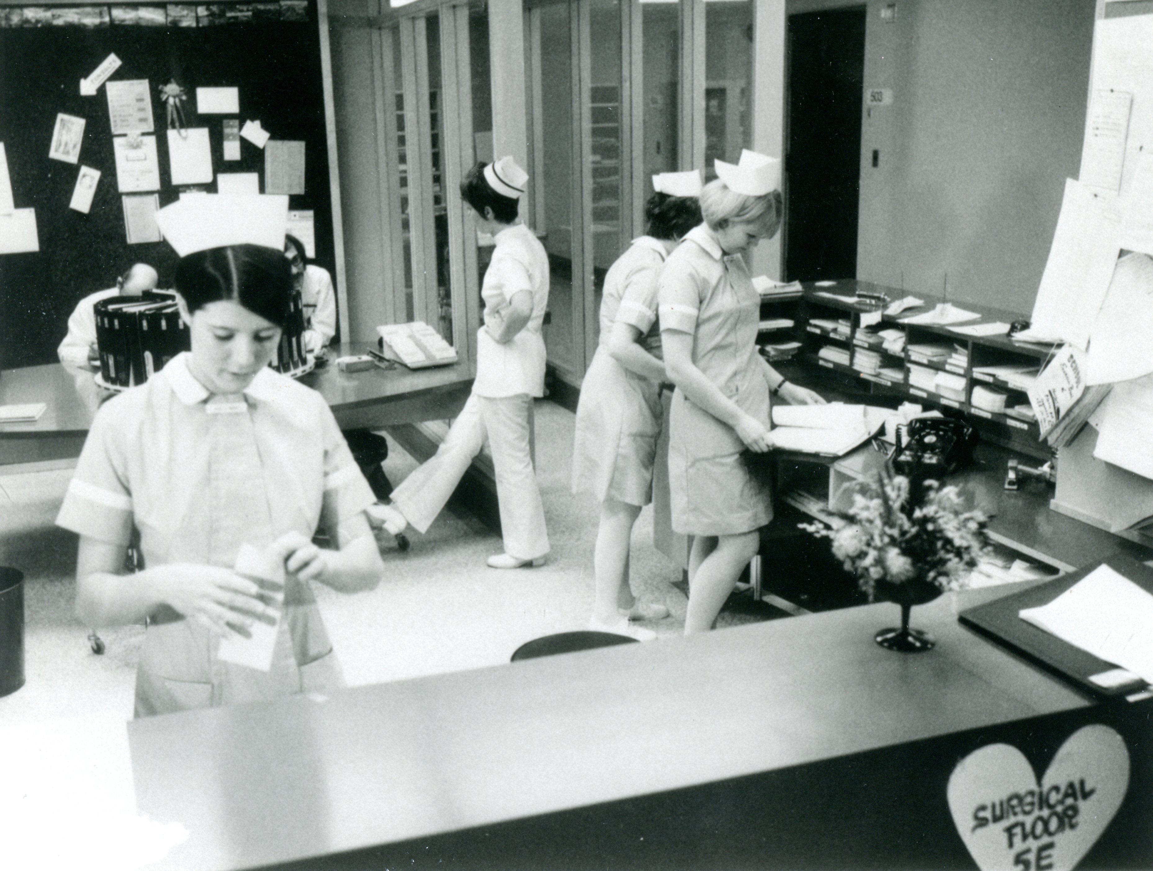 Six people at a reception area are talking and handling papers. Four women are wearing nurses' caps.