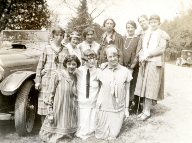 Ten women dressed casually pose beside a motor vehicle on a sunny day. There are trees in the background.