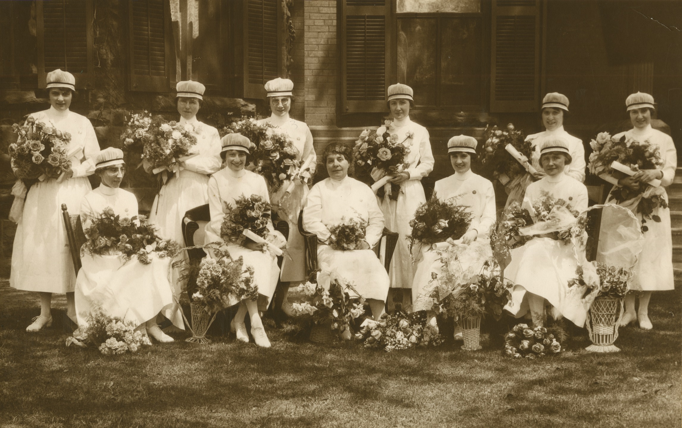 Eleven smiling women pose formally with flower arrangements. All but one are wearing nurses' caps.