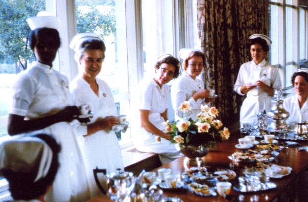 Six young women dressed in nurses uniforms stand in a room with large windows before a decorated table filled with tea and cookies.