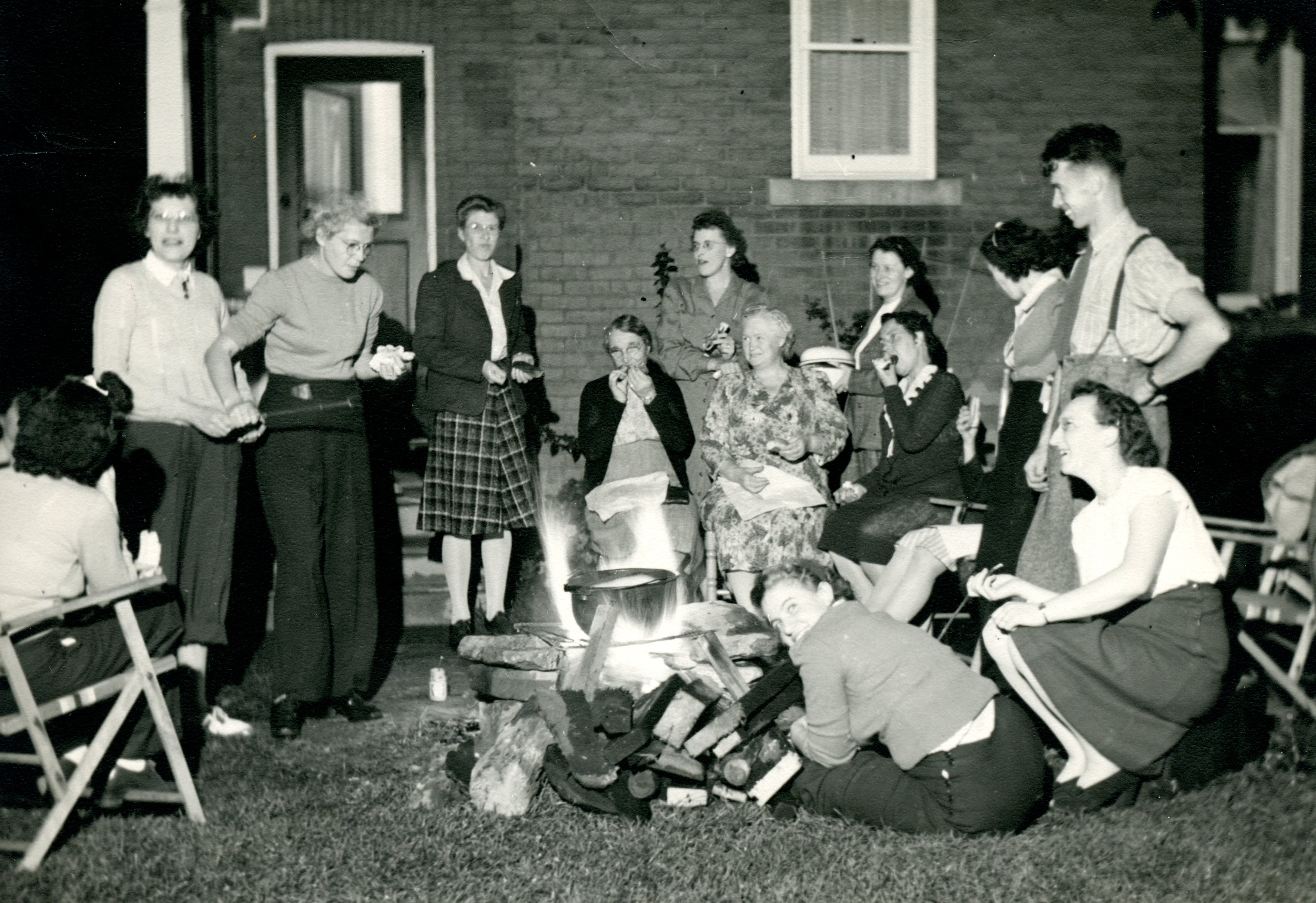 Thirteen people gather around a fire, eating corn. They are in front of a brick building.