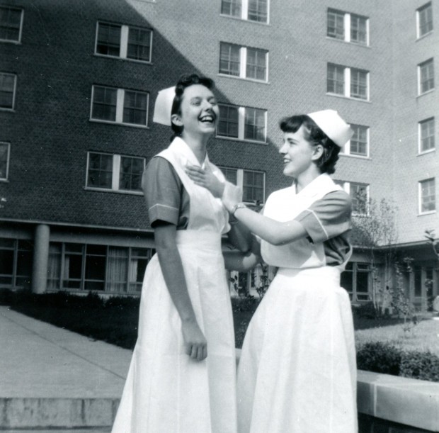 Two young women dressed in nurses' uniforms and caps joke affectionately before a tall brick building.