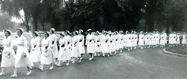 Over 30 women march in a row, wearing white uniforms, caps, and shoes.